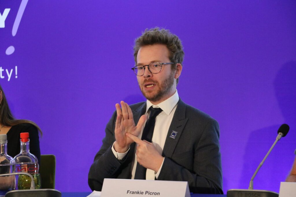 A man in a suit and glasses speaks at a panel, gesturing with his hands, with a nameplate reading "Frankie Picron" in front of him against a purple background.