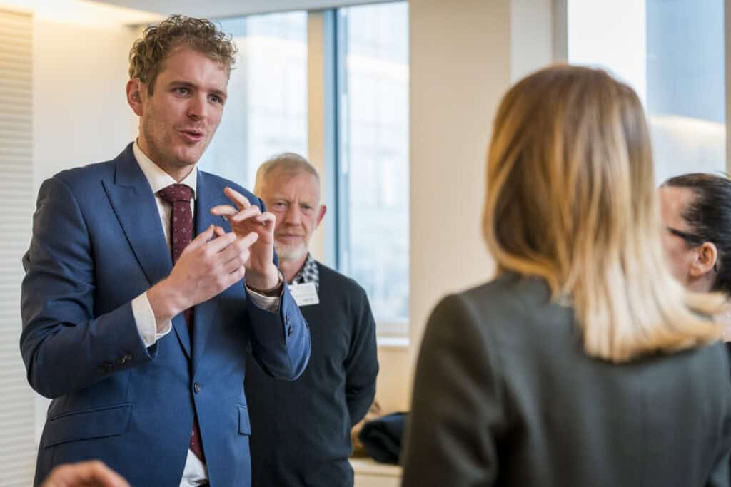 A man in a blue suit speaks to a small group of people in an office setting, gesturing with his hands as others listen attentively.