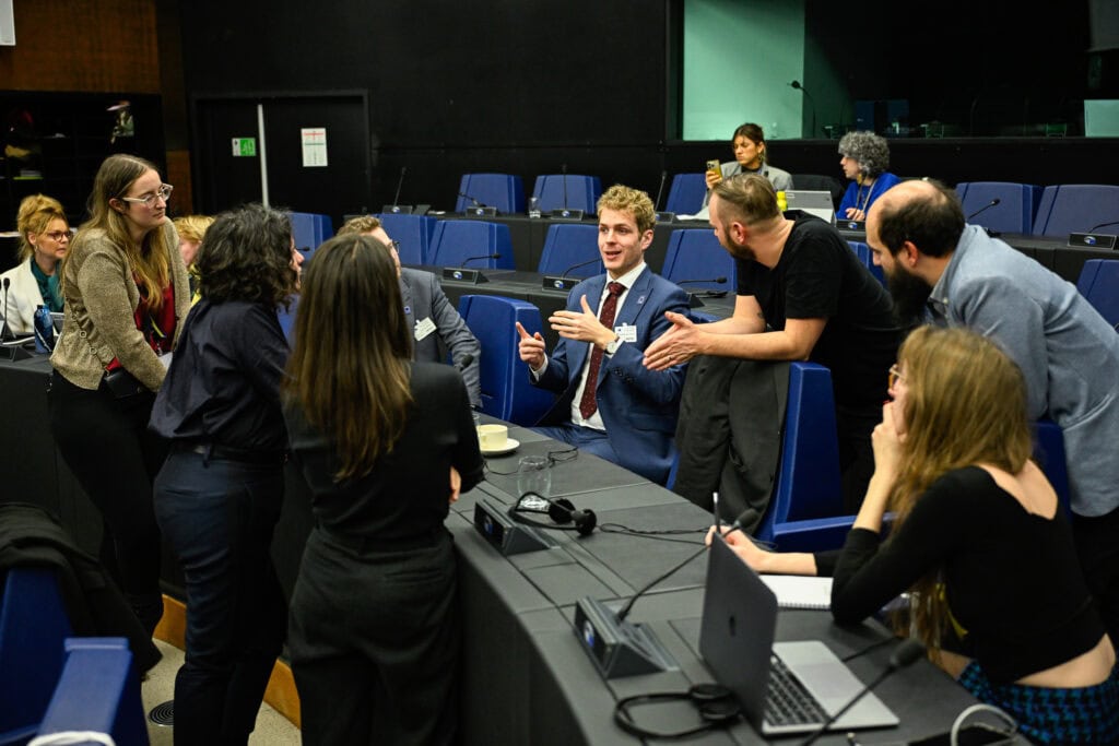 A group of people, some seated and some standing, engage in a discussion around conference tables in a modern meeting room.