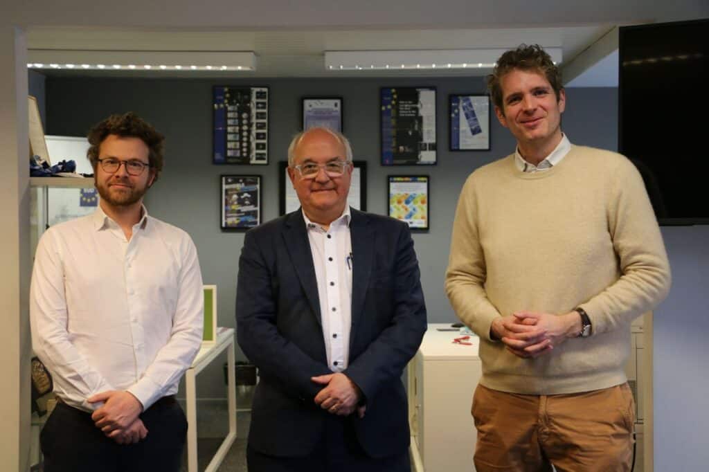 Three men stand side by side in an office setting, dressed in business casual attire, with framed documents and monitors on the wall behind them.