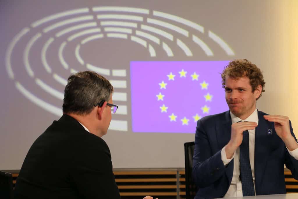Two men in suits have a discussion in front of a screen displaying the European Union flag and the European Parliament logo.