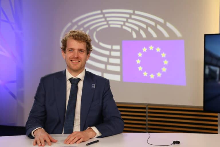 A man in a blue suit sits at a desk with the EU flag and European Parliament emblem projected on the wall behind him.