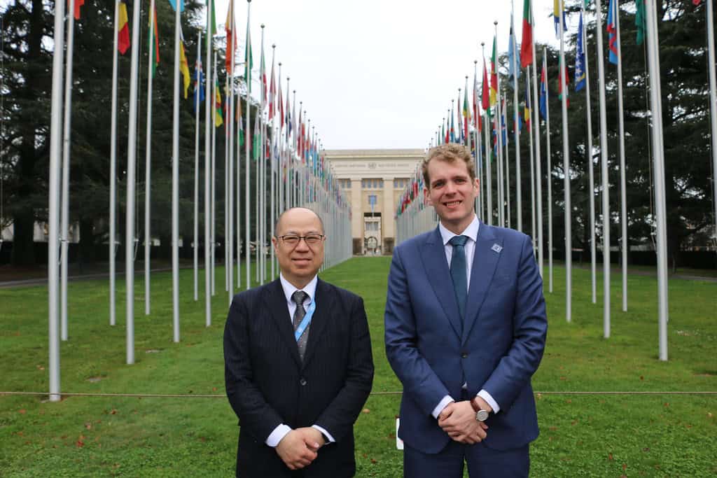 Two men in suits stand on a lawn flanked by flagpoles with international flags, with a large building in the background.