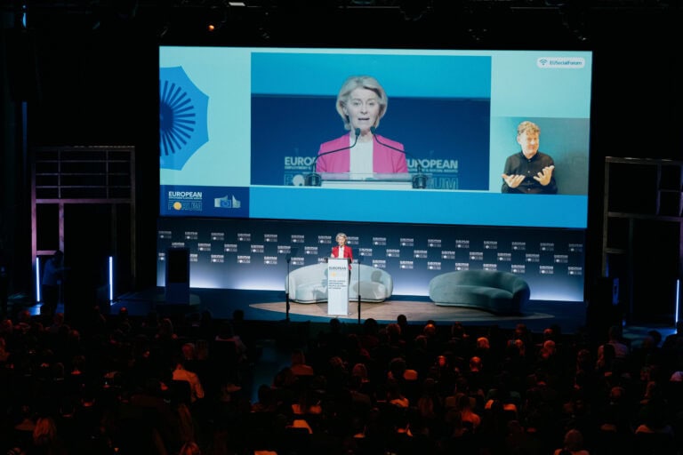 A woman speaks at a lectern on stage at a European Commission event, with her image projected on a large screen behind her and an interpreter visible at the side.
