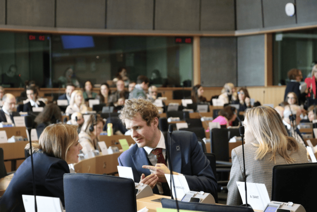 People seated in a large conference room, with three individuals in the foreground engaged in conversation. Nameplates are visible on tables, and a group of attendees sits in the background.