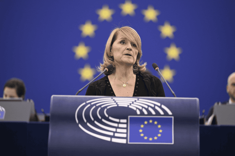 A woman speaks at a lectern with the European Union emblem, with the EU flag and yellow stars visible in the background.