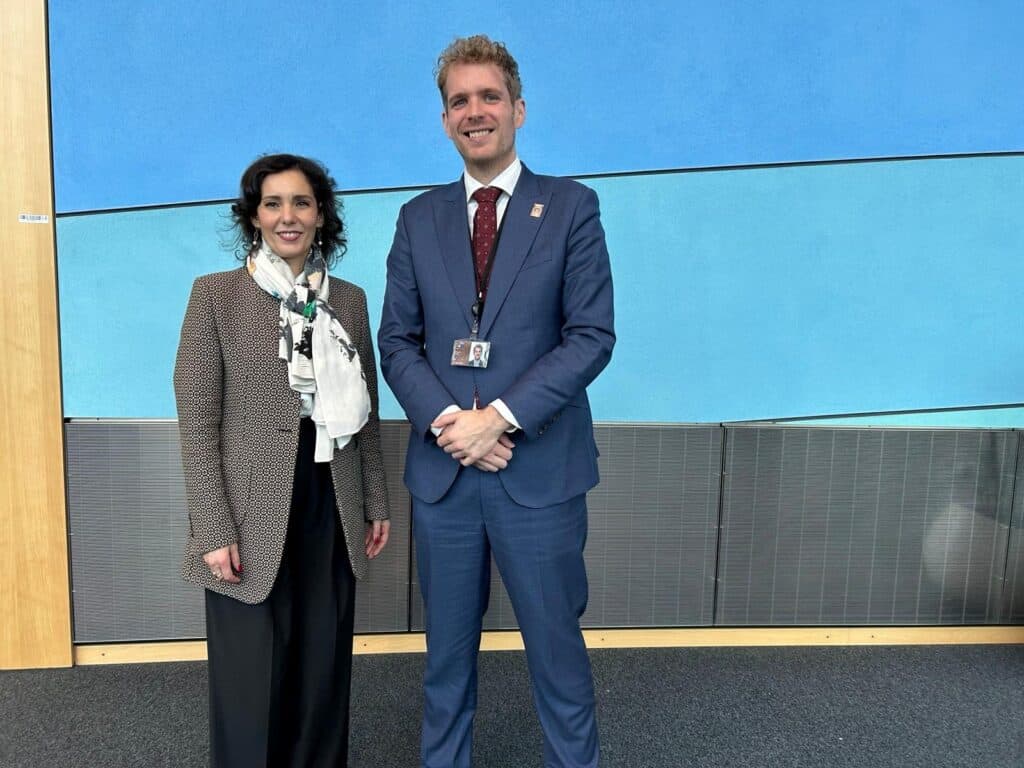 Two people, one woman and one man in business attire, stand side by side in front of a blue wall indoors.