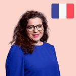 A woman with curly brown hair and glasses wears a blue top, standing in front of a pink background with a French flag in the top right-hand corner.