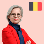 A woman with short grey hair and glasses wearing a red blazer poses with arms folded; a Belgian flag appears in the top right corner.
