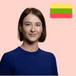 A woman with straight brown hair in a blue top stands in front of a pink background with the Lithuanian flag in the top right-hand corner.