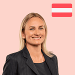 A woman in a black blazer smiles in front of a pink background with the Austrian flag in the top right-hand corner.