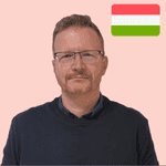 A man with short fair hair and glasses wearing a dark jumper stands in front of a light pink background with a Hungarian flag in the top right corner.