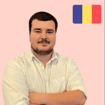 A man with short dark hair and a beard, wearing a white shirt, stands with arms folded in front of a pink background with a Romanian flag in the top right corner.