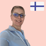 Smiling person with short hair and glasses stands in front of a light pink background with a Finnish flag in the top right-hand corner.