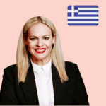 A woman with straight blonde hair in a black blazer and white shirt smiles in front of a light pink background with a Greek flag in the top right-hand corner.