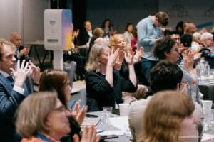 People seated at tables in a conference room clap their hands, some with drinks and papers in front of them. A few individuals stand or look at their mobiles in the background.